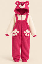 A person wearing a plush teddy bear onesie pajama with a hood, featuring a pink body, cream sleeves, and paw-print details on the legs, posed indoors against a beige wall.