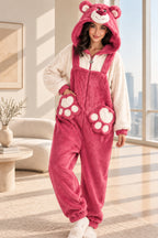 A person wearing a plush teddy bear onesie pajama with a hood, featuring a pink body, cream sleeves, and paw-print details on the legs, posed indoors against a beige wall.