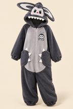 Woman wearing matching dark gray plush bunny onesies in a living room setting. The hoods feature long bunny ears, a cartoon face with white teeth, and paw details on the front.