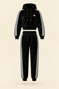 A black Adidas tracksuit laid flat on a white fabric background, featuring a velour hoodie with white three-stripe detailing on the sleeves and chest logo, paired with matching black jogger pants.