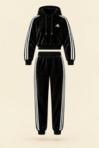 A black Adidas tracksuit laid flat on a white fabric background, featuring a velour hoodie with white three-stripe detailing on the sleeves and chest logo, paired with matching black jogger pants.