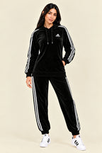 A black Adidas tracksuit laid flat on a white fabric background, featuring a velour hoodie with white three-stripe detailing on the sleeves and chest logo, paired with matching black jogger pants.