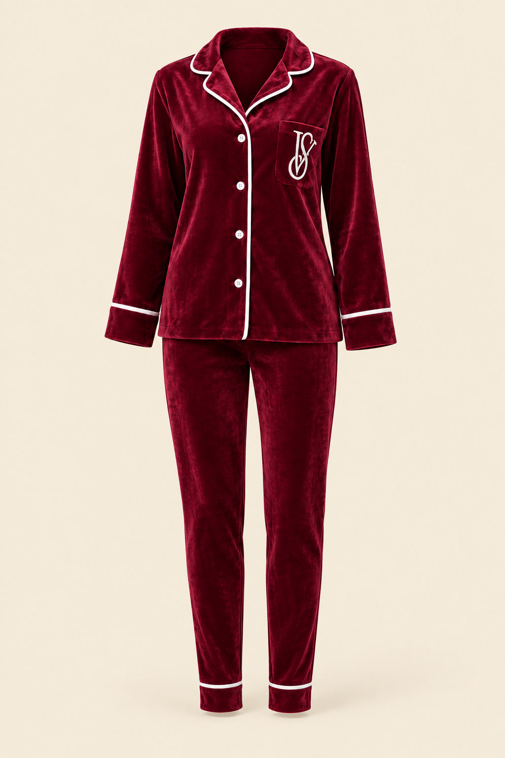 A woman wearing a matching burgundy velvet pajama set with white piping and a white embroidered monogram, sitting on a pink tufted sofa in an indoor setting.