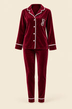A woman wearing a matching burgundy velvet pajama set with white piping and a white embroidered monogram, sitting on a pink tufted sofa in an indoor setting.