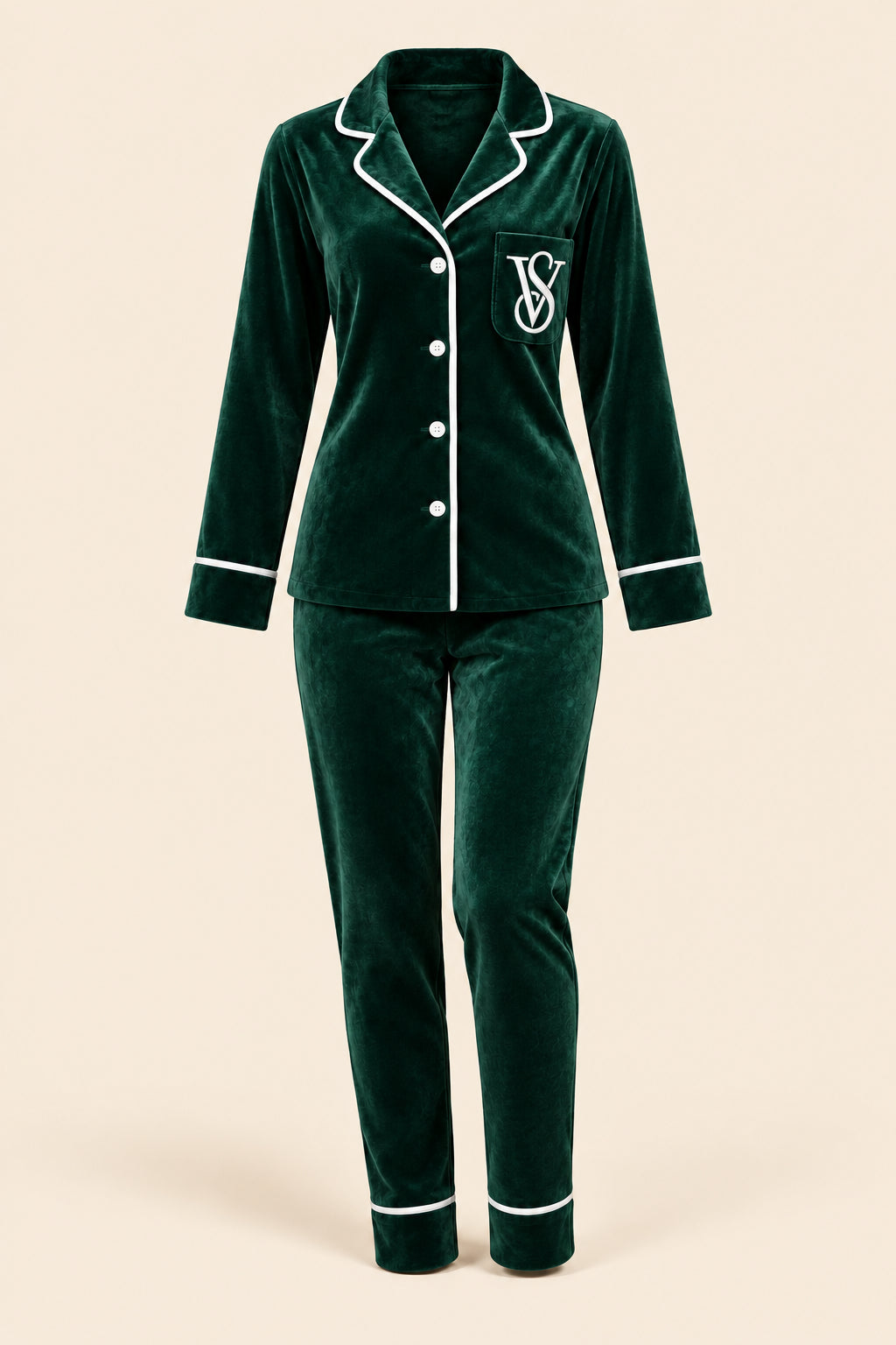 A woman wearing a dark green velvet pajama set with white piping and a white embroidered monogram logo on the chest, sitting on a pink velvet sofa in an indoor setting.