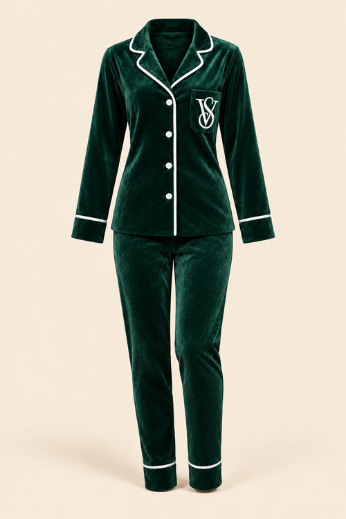 A woman wearing a dark green velvet pajama set with white piping and a white embroidered monogram logo on the chest, sitting on a pink velvet sofa in an indoor setting.