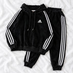 A black Adidas tracksuit laid flat on a white fabric background, featuring a velour hoodie with white three-stripe detailing on the sleeves and chest logo, paired with matching black jogger pants.