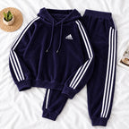 A dark purple Adidas tracksuit laid flat on a white fabric background, featuring a hooded sweatshirt with white three-stripes and a matching pair of jogger pants. A small white Adidas logo is visible on the chest.
