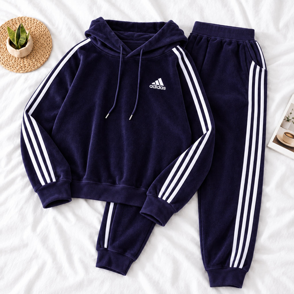 A dark purple Adidas tracksuit laid flat on a white fabric background, featuring a hooded sweatshirt with white three-stripes and a matching pair of jogger pants. A small white Adidas logo is visible on the chest.