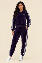 A two-piece Adidas tracksuit in deep purple velvet, featuring a hooded sweatshirt with white three-stripes on the sleeves and a white Adidas logo on the chest, paired with matching jogger pants with white three-stripes down the side, shown against a solid light beige background.