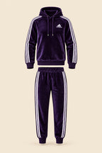 A dark purple Adidas tracksuit laid flat on a white fabric background, featuring a hooded sweatshirt with white three-stripes and a matching pair of jogger pants. A small white Adidas logo is visible on the chest.