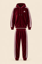 A maroon Adidas tracksuit set consisting of a hooded sweatshirt and matching joggers, laid flat on a white fabric background. The tracksuit features white three-stripes on the sleeves and legs, a white Adidas logo on the chest, and white drawstrings.