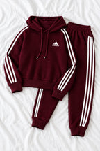 A maroon Adidas tracksuit set consisting of a hooded sweatshirt and matching joggers, laid flat on a white fabric background. The tracksuit features white three-stripes on the sleeves and legs, a white Adidas logo on the chest, and white drawstrings.