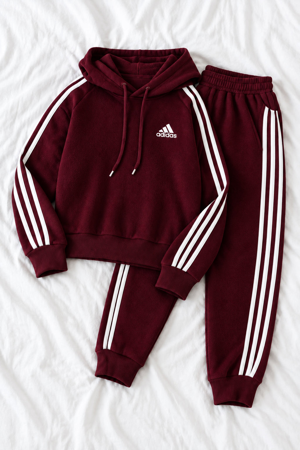 A maroon Adidas tracksuit set consisting of a hooded sweatshirt and matching joggers, laid flat on a white fabric background. The tracksuit features white three-stripes on the sleeves and legs, a white Adidas logo on the chest, and white drawstrings.