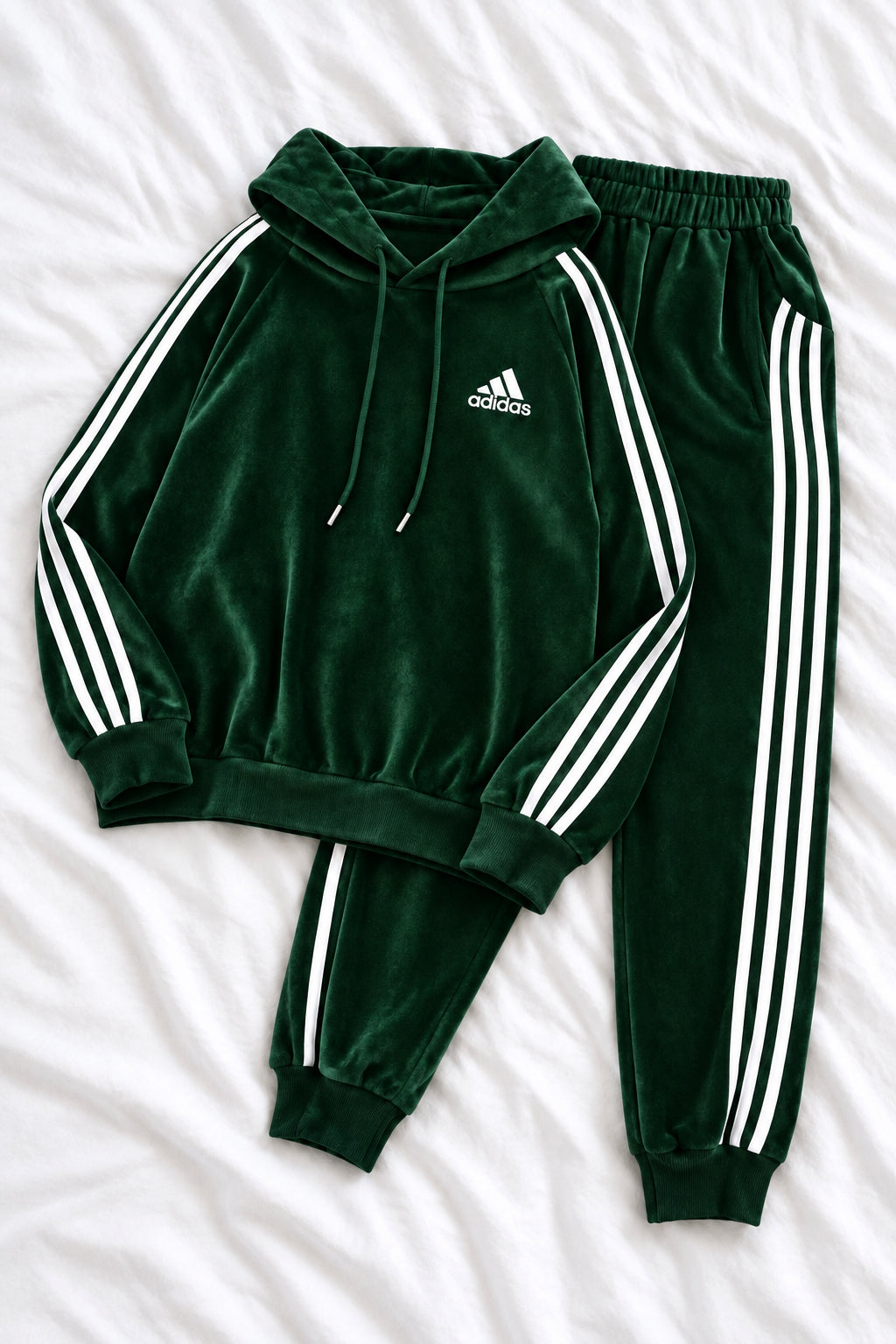 A dark green Adidas tracksuit laid flat on a white fabric background, featuring a hooded sweatshirt with white three-stripe detailing on the sleeves and a matching pair of jogger pants.