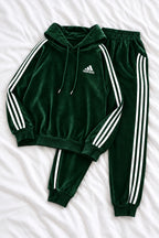 A dark green Adidas tracksuit laid flat on a white fabric background, featuring a hooded sweatshirt with white three-stripe detailing on the sleeves and a matching pair of jogger pants.