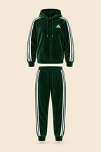 A dark green Adidas tracksuit laid flat on a white fabric background, featuring a hooded sweatshirt with white three-stripe detailing on the sleeves and a matching pair of jogger pants.