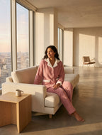 A woman wearing a pink plush pajama set with white trim sits barefoot on a cream sofa in a sunlit modern loft with large windows overlooking a city skyline.