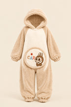 A woman wearing a beige and cream plush hooded pajama set with a cartoon rabbit graphic on the front, sitting on a fluffy rug and making a peace sign. A rabbit plush toy sits on a box to her left, and a brand logo overlay for 'Hesham Lotte' is in the top-left corner.
