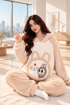 A woman wearing a beige and cream plush hooded pajama set with a cartoon rabbit graphic on the front, sitting on a fluffy rug and making a peace sign. A rabbit plush toy sits on a box to her left, and a brand logo overlay for 'Hesham Lotte' is in the top-left corner.