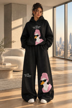 A woman modeling a black two-piece lounge set with a pink and white duck graphic and white text, standing in a room with a dark paneled wall and gray floor. The image includes 'Rose home wear' branding and a pink 'ROSE' banner on the left.