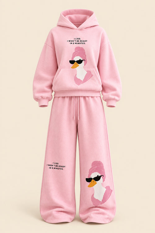 A woman modeling a pink two-piece loungewear set consisting of a pullover hoodie and matching wide-leg sweatpants. The set features a cartoon duck graphic wearing sunglasses on the front of both pieces, with small black text printed on the chest and legs.