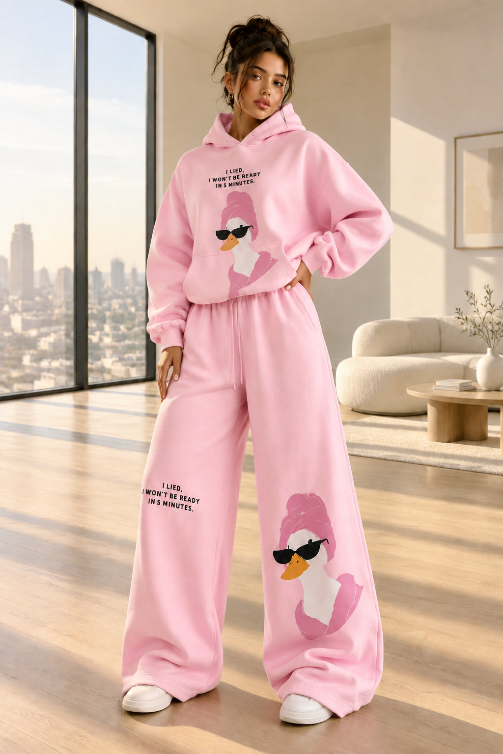 A woman modeling a pink two-piece loungewear set consisting of a pullover hoodie and matching wide-leg sweatpants. The set features a cartoon duck graphic wearing sunglasses on the front of both pieces, with small black text printed on the chest and legs.