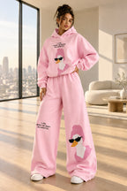 A woman modeling a pink two-piece loungewear set consisting of a pullover hoodie and matching wide-leg sweatpants. The set features a cartoon duck graphic wearing sunglasses on the front of both pieces, with small black text printed on the chest and legs.