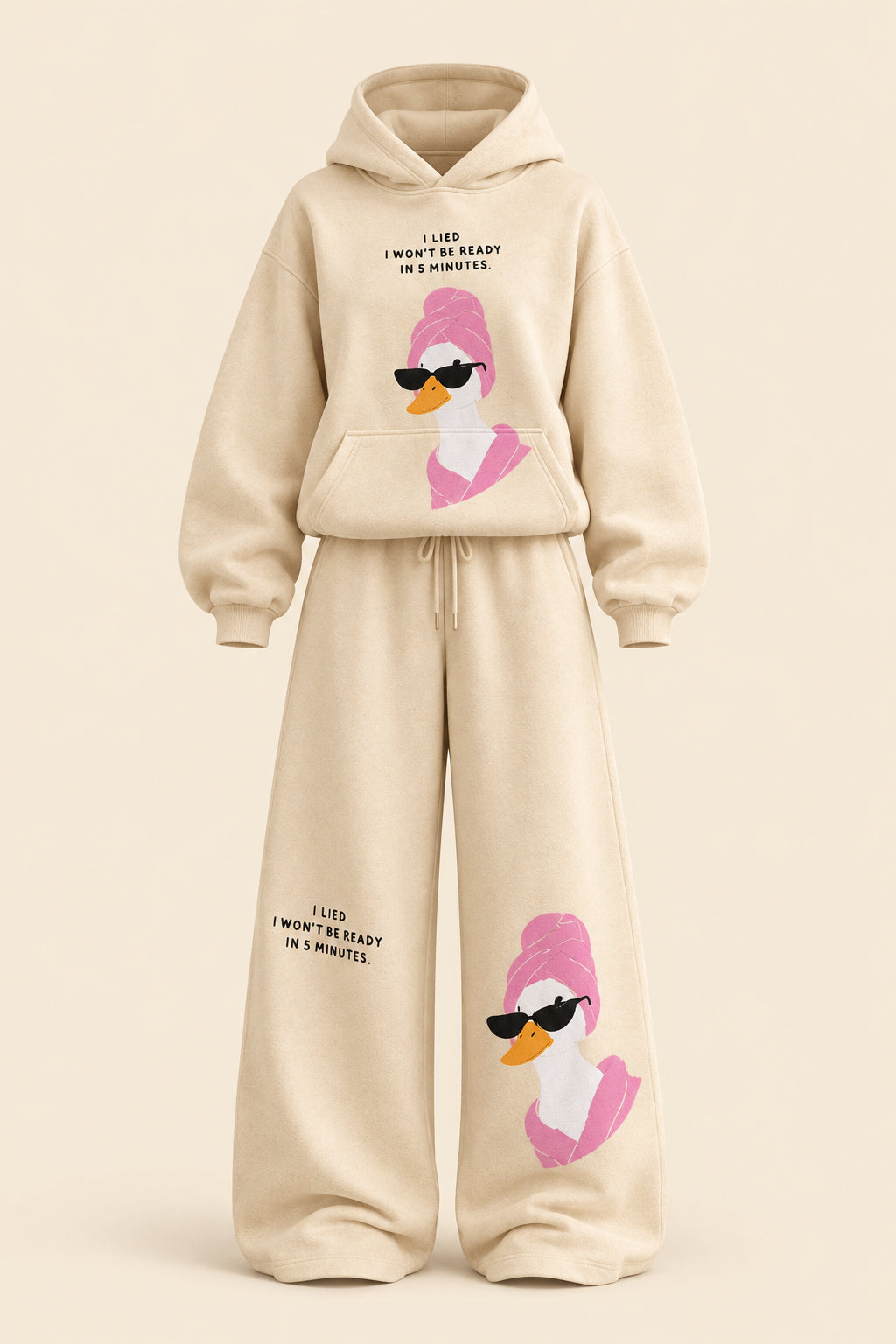 A woman modeling a beige two-piece lounge set with a matching hoodie and wide-leg sweatpants, both featuring a pink cartoon duck graphic with black sunglasses and small text. The background is a dark paneled wall with gray floor and a gray bench.