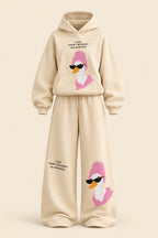 A woman modeling a beige two-piece lounge set with a matching hoodie and wide-leg sweatpants, both featuring a pink cartoon duck graphic with black sunglasses and small text. The background is a dark paneled wall with gray floor and a gray bench.