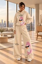 A woman modeling a beige two-piece lounge set with a matching hoodie and wide-leg sweatpants, both featuring a pink cartoon duck graphic with black sunglasses and small text. The background is a dark paneled wall with gray floor and a gray bench.