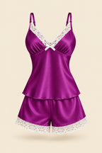 A woman models a two-piece satin pajama set in a luxurious purple shade with white lace trim. The set includes a camisole top and matching shorts.