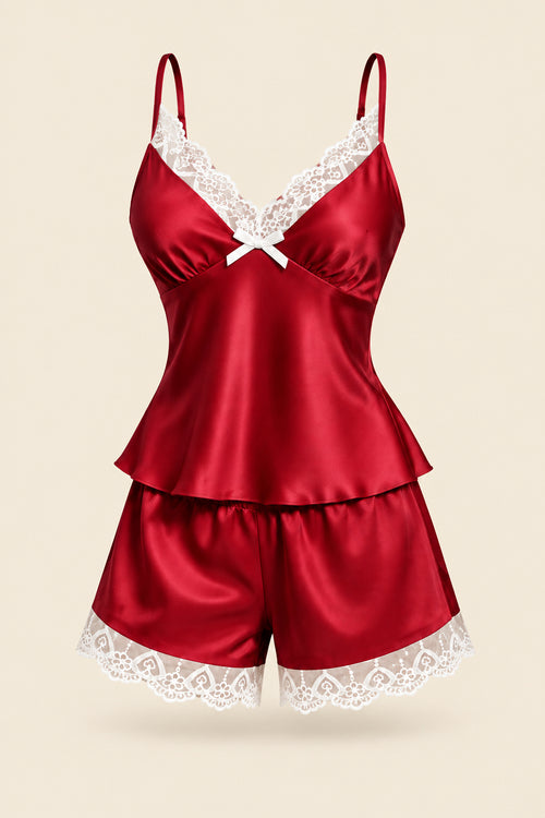A woman modeling a satin sleepwear set in a dimly lit bathroom setting. The set consists of a deep red camisole top with white lace trim and matching red lace-trimmed shorts.