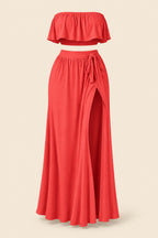 A woman models a coral-red two-piece lingerie set in a furnished indoor room. The set includes a strapless ruffled crop top and a high-slit maxi skirt.