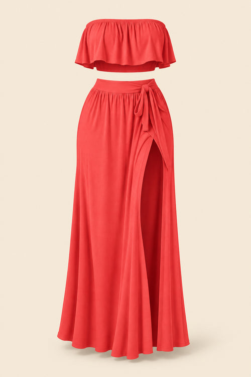 A woman models a coral-red two-piece lingerie set in a furnished indoor room. The set includes a strapless ruffled crop top and a high-slit maxi skirt.