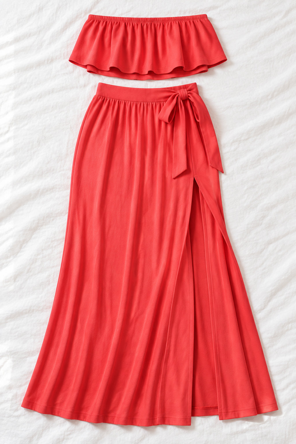 A woman models a coral-red two-piece lingerie set in a furnished indoor room. The set includes a strapless ruffled crop top and a high-slit maxi skirt.