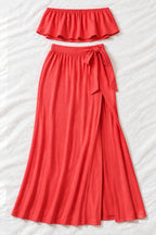 A woman models a coral-red two-piece lingerie set in a furnished indoor room. The set includes a strapless ruffled crop top and a high-slit maxi skirt.