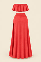 A woman models a coral-red two-piece lingerie set in a furnished indoor room. The set includes a strapless ruffled crop top and a high-slit maxi skirt.