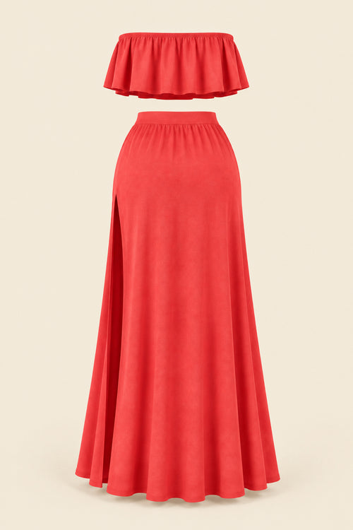 A woman models a coral-red two-piece lingerie set in a furnished indoor room. The set includes a strapless ruffled crop top and a high-slit maxi skirt.