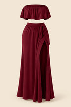 A woman modeling a burgundy two-piece lingerie set in a luxurious indoor setting. The set includes a strapless ruffled crop top and a matching high-slit maxi skirt.