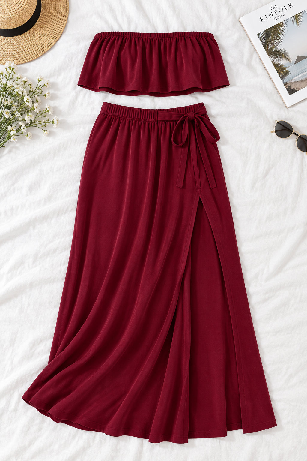 A woman modeling a burgundy two-piece lingerie set in a luxurious indoor setting. The set includes a strapless ruffled crop top and a matching high-slit maxi skirt.