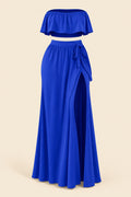 A woman modeling a royal blue two-piece lingerie set in a luxurious indoor setting. The set includes a strapless ruffled crop top and a matching high-slit maxi skirt.