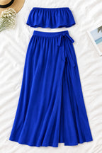 A woman modeling a royal blue two-piece lingerie set in a luxurious indoor setting. The set includes a strapless ruffled crop top and a matching high-slit maxi skirt.