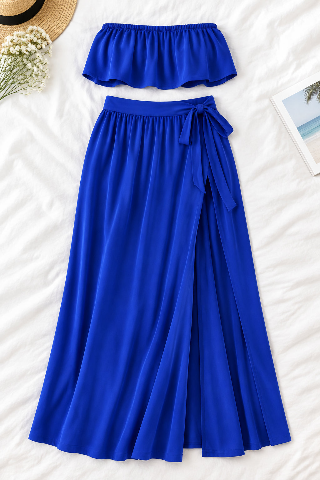 A woman modeling a royal blue two-piece lingerie set in a luxurious indoor setting. The set includes a strapless ruffled crop top and a matching high-slit maxi skirt.
