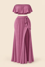 A woman modeling a mauve two-piece lingerie set in a furnished indoor room, with a smaller inset view showing the back of the set. The set includes a strapless ruffled crop top and a high-slit maxi skirt.