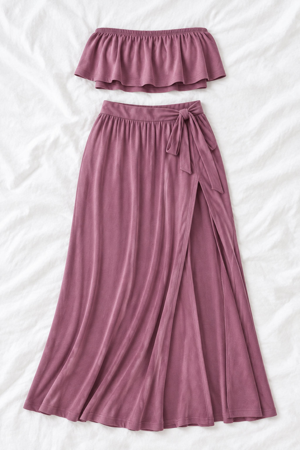 A woman modeling a mauve two-piece lingerie set in a furnished indoor room, with a smaller inset view showing the back of the set. The set includes a strapless ruffled crop top and a high-slit maxi skirt.