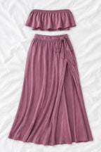 A woman modeling a mauve two-piece lingerie set in a furnished indoor room, with a smaller inset view showing the back of the set. The set includes a strapless ruffled crop top and a high-slit maxi skirt.