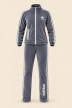 A woman modeling a gray Adidas tracksuit consisting of a zip-up jacket and matching pants, with white Adidas logos on the chest and down the sides. The image has a 'Magic Queen' watermark in the top left, a phone number on the right, and a model badge at the bottom.
