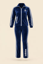 A woman modeling a navy blue Adidas tracksuit with white stripes and logos, standing outdoors on a paved walkway with trees in the background. The image includes 'Magic Queen' branding and contact details.