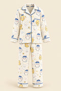 A woman takes a mirror selfie wearing a two-piece cartoon pajama set with a cream base and blue, yellow, and pink printed characters. She is sitting on a beige couch with plush toys in the background.